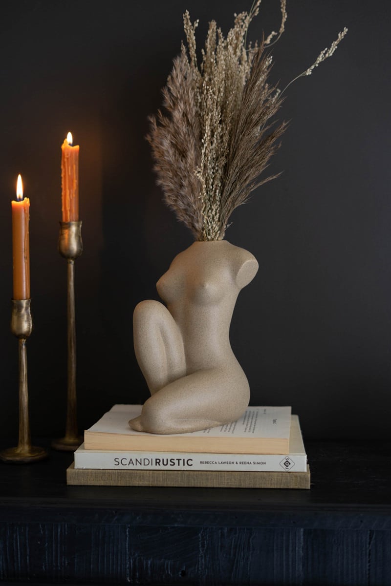 Beautiful half kneeling nude female vase with a sprig branch coming out of its neck. The vase is cream with two taper candles lit behind it. Fine luxurious blue background and three books underneath.